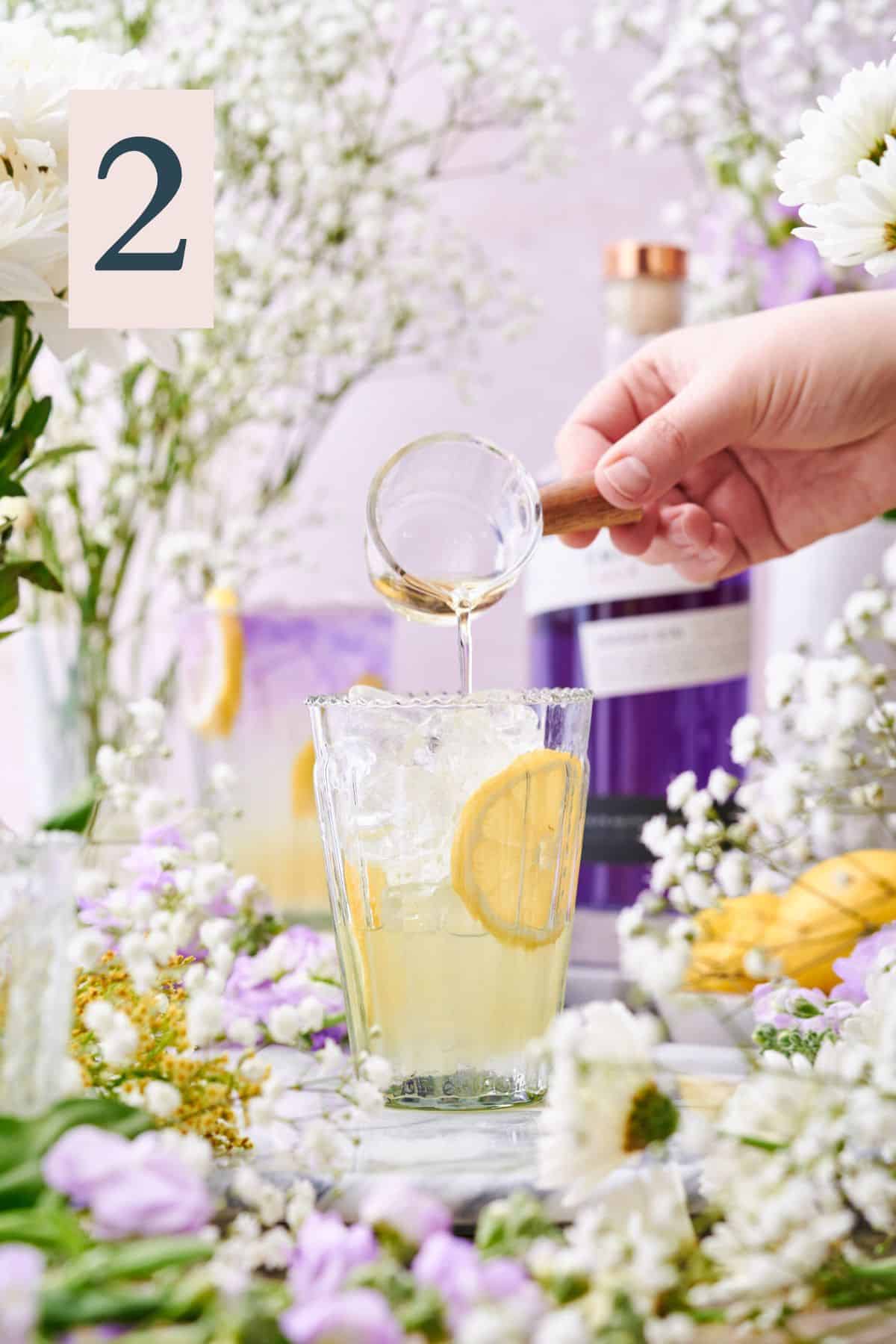 hand pouring liqueur into a glass with lemon wheels and ice surrounded by lots of white and purple flowers.