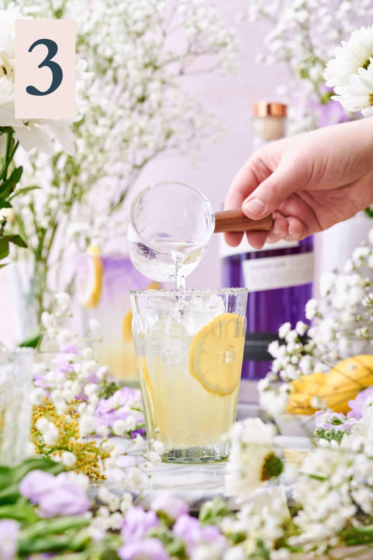 hand pouring soda water into a glass with lemon wheels and ice surrounded by lots of white and purple flowers. 