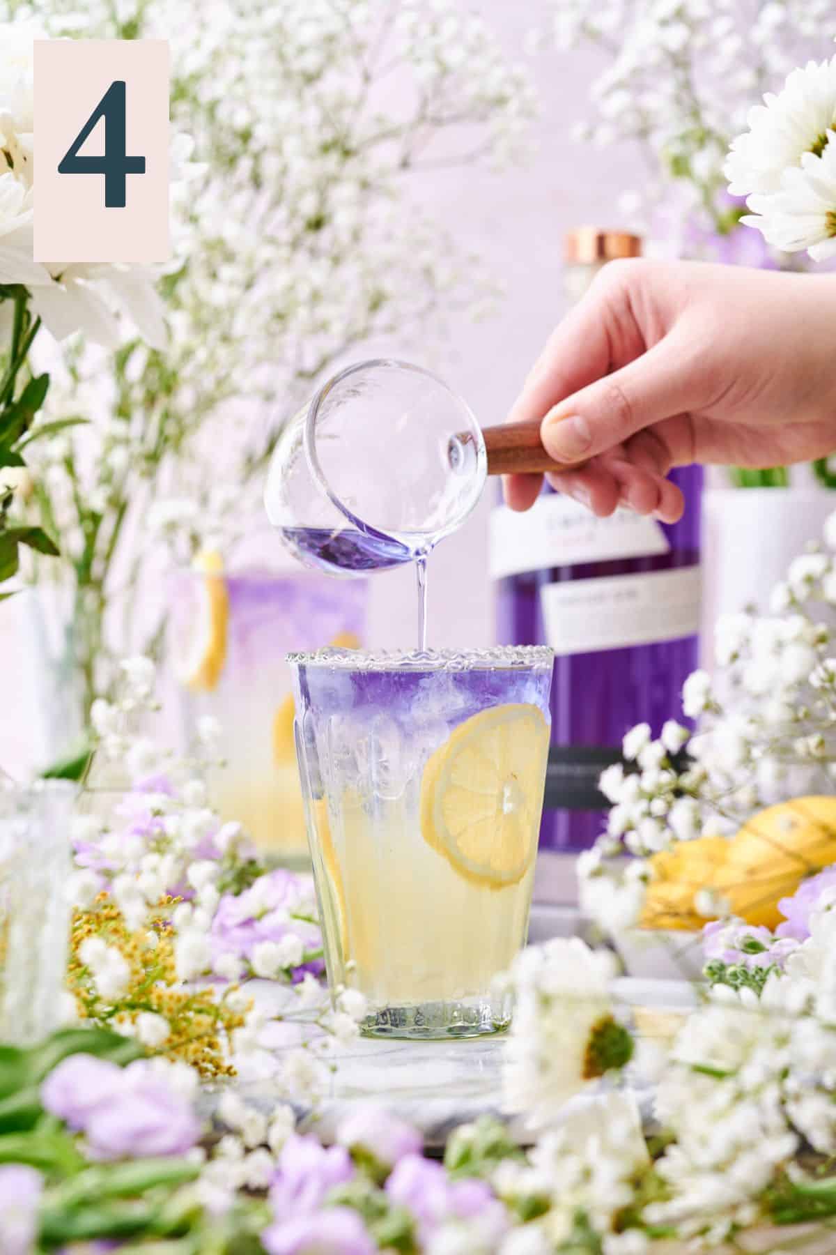 hand pouring purple Empress gin into a glass with lemon wheels and ice surrounded by lots of white and purple flowers.
