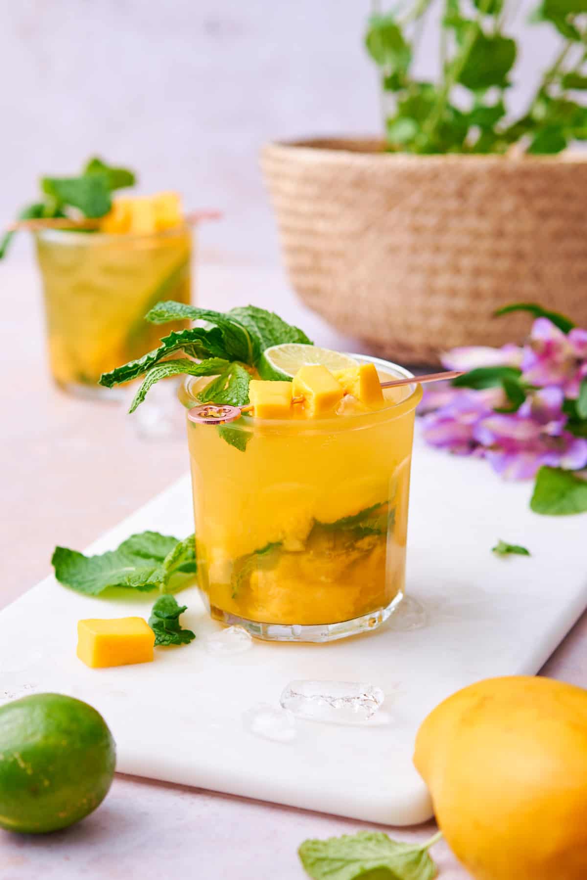 Mango Mojito cocktail with lime and mint garnish on top, surrounded by flowers, another cocktail and a light warm background.
