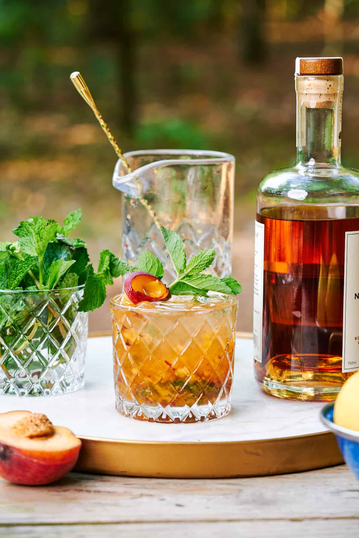 peach bourbon smash with fresh mint, peaches, and a mixing glass on a white marble tray with a forest in the background.