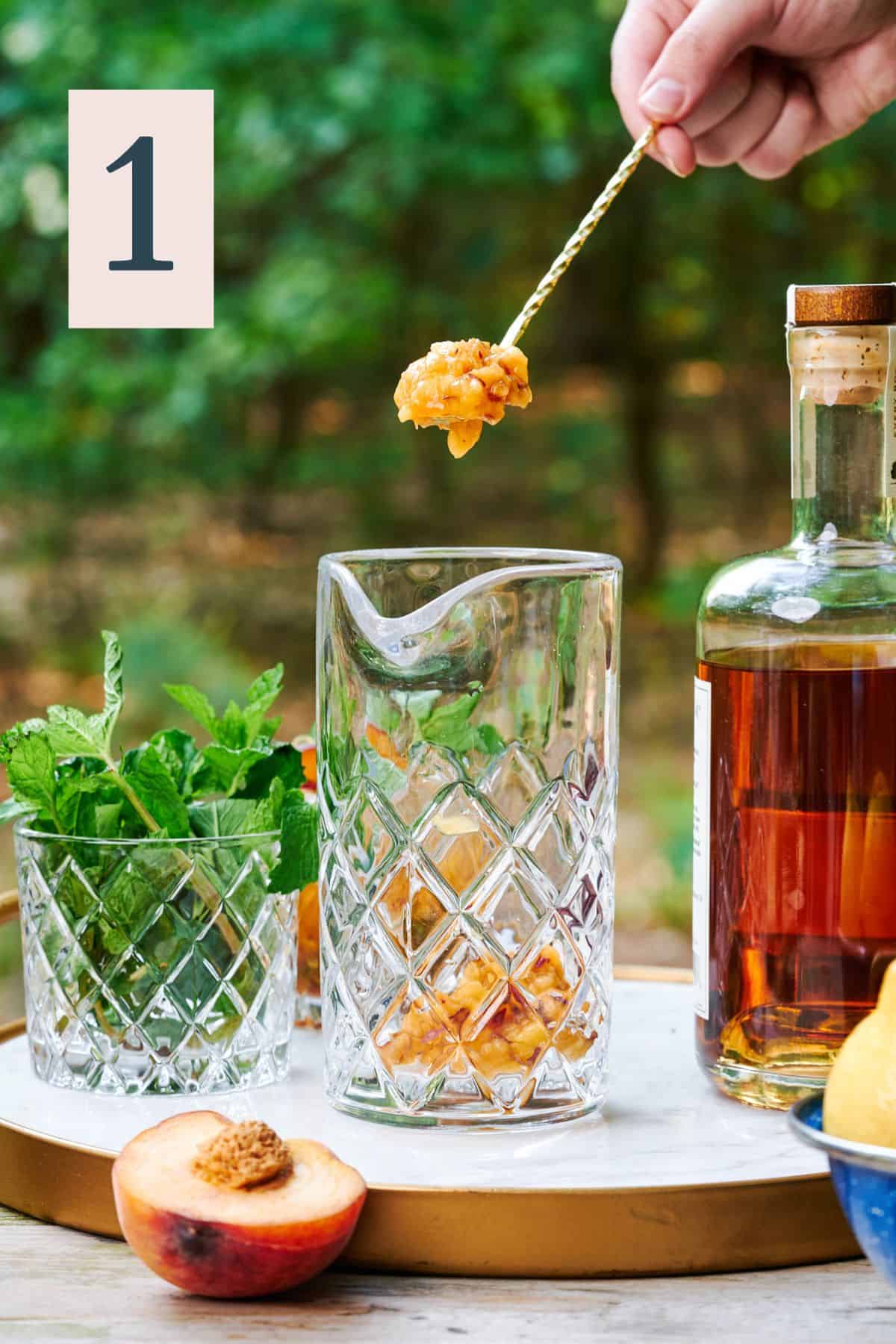 hand holding some small peach bits over top a mixing glass in the woods with bourbon and fresh mint on the scene.