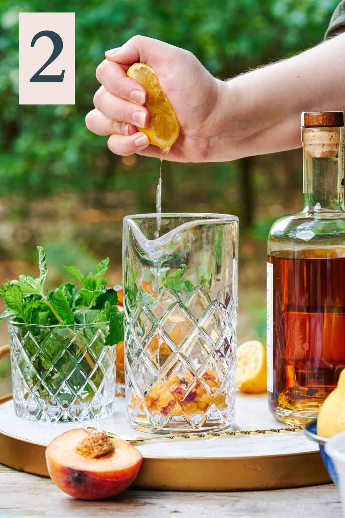 hand squeezing lemon juice over top of a mixing glass in the woods with bourbon and fresh mint on the scene.