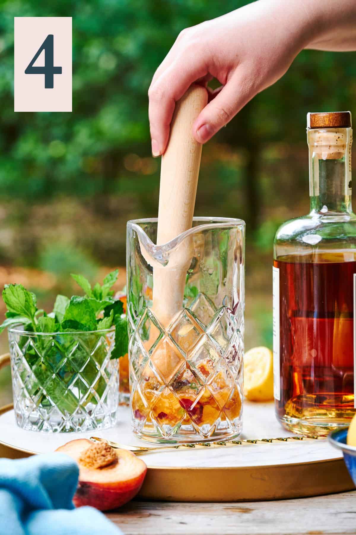 hand using a wooden muddler to combine the mint into a drink with peaches in a glass mixing glass in the woods. 