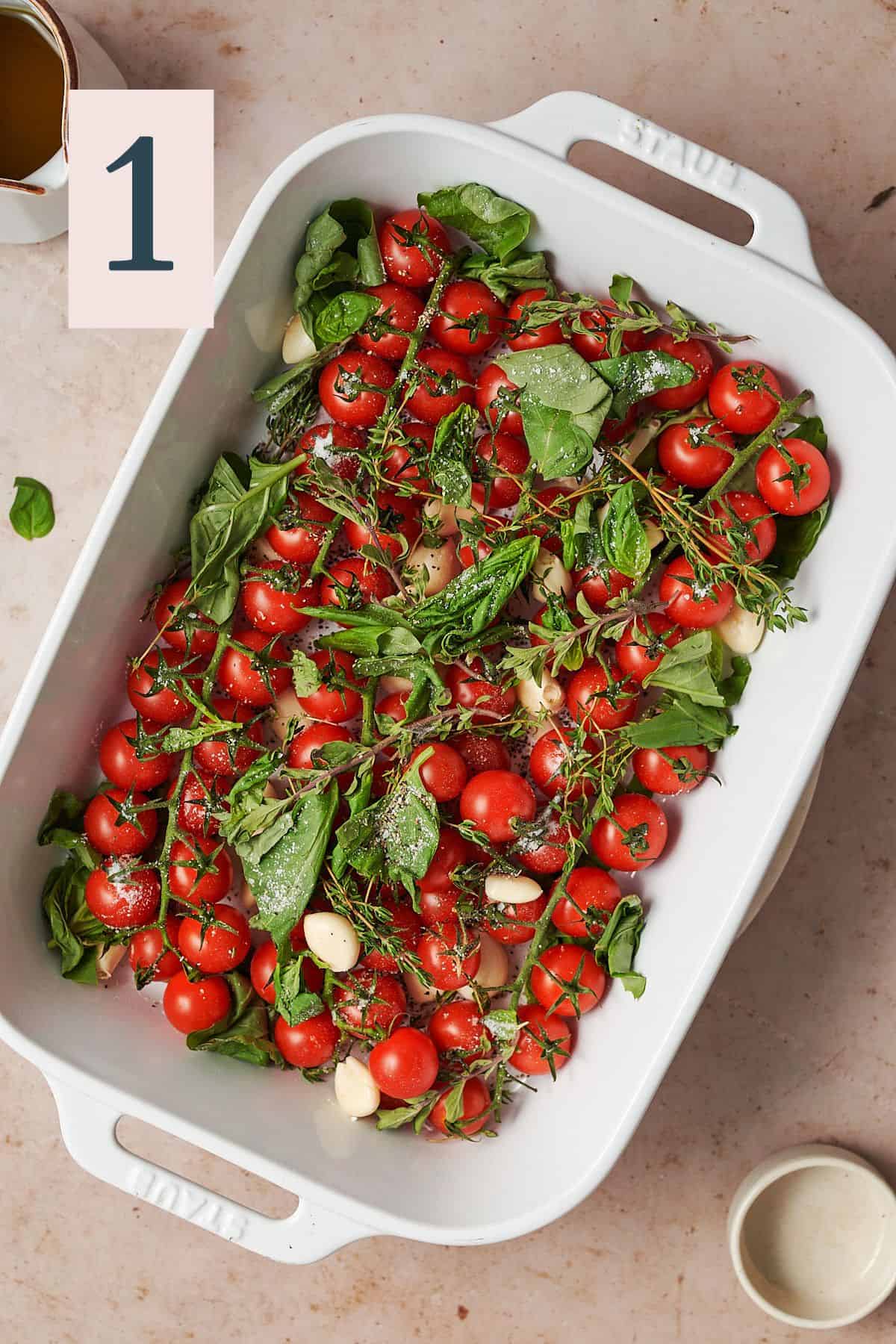 cherry tomatoes in a white baking dish topped with herbs, salt, pepper, and olive oil. 