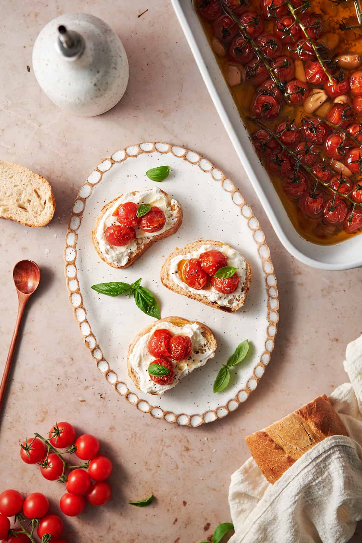 toasts with whipped ricotta and cherry tomato confit and fresh basil on top, with a large baking dish full of more tomato confit on the side. 