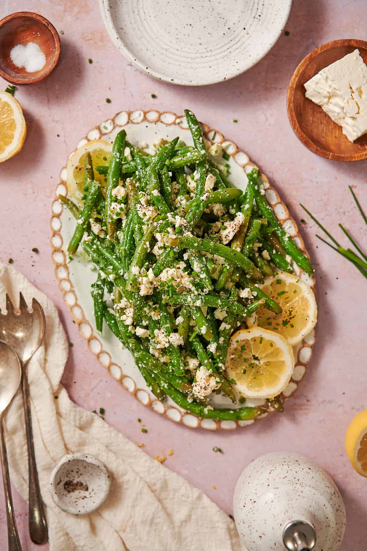 a plate of green bean feta salad surrounded by lemon slices, chives, salt, and pepper. 