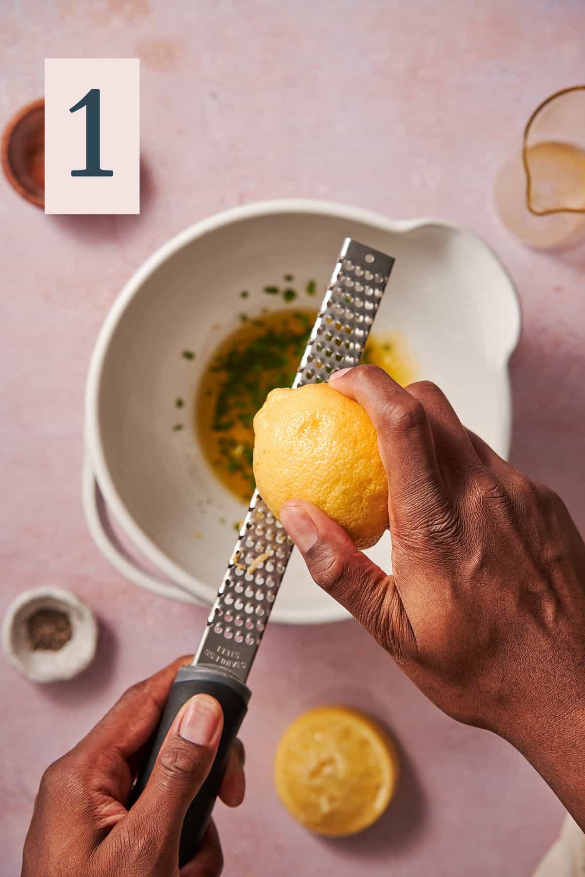 hand zesting a lemon in a bowl with herbs and olive oil. 