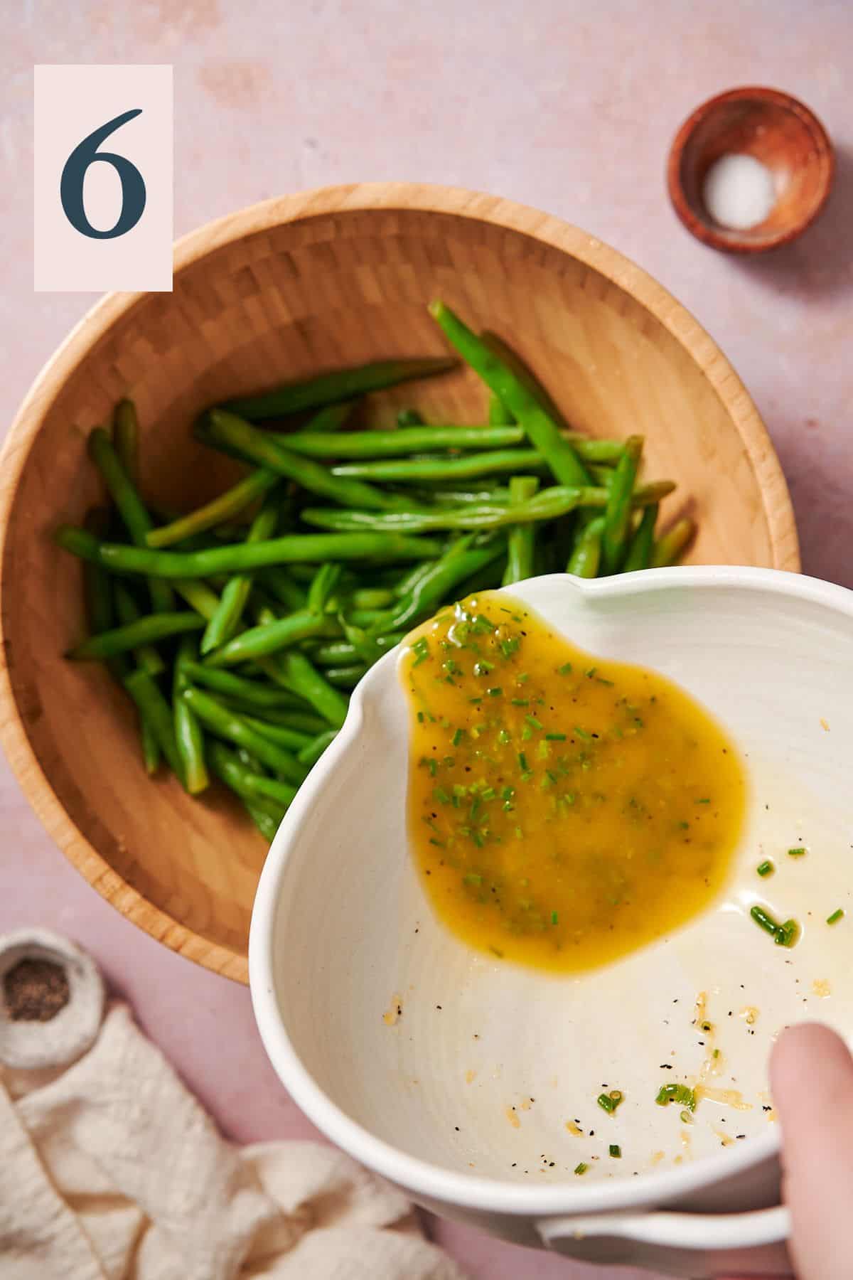 pouring lemon olive oil dressing with chives into a bowl of cooked green beans. 