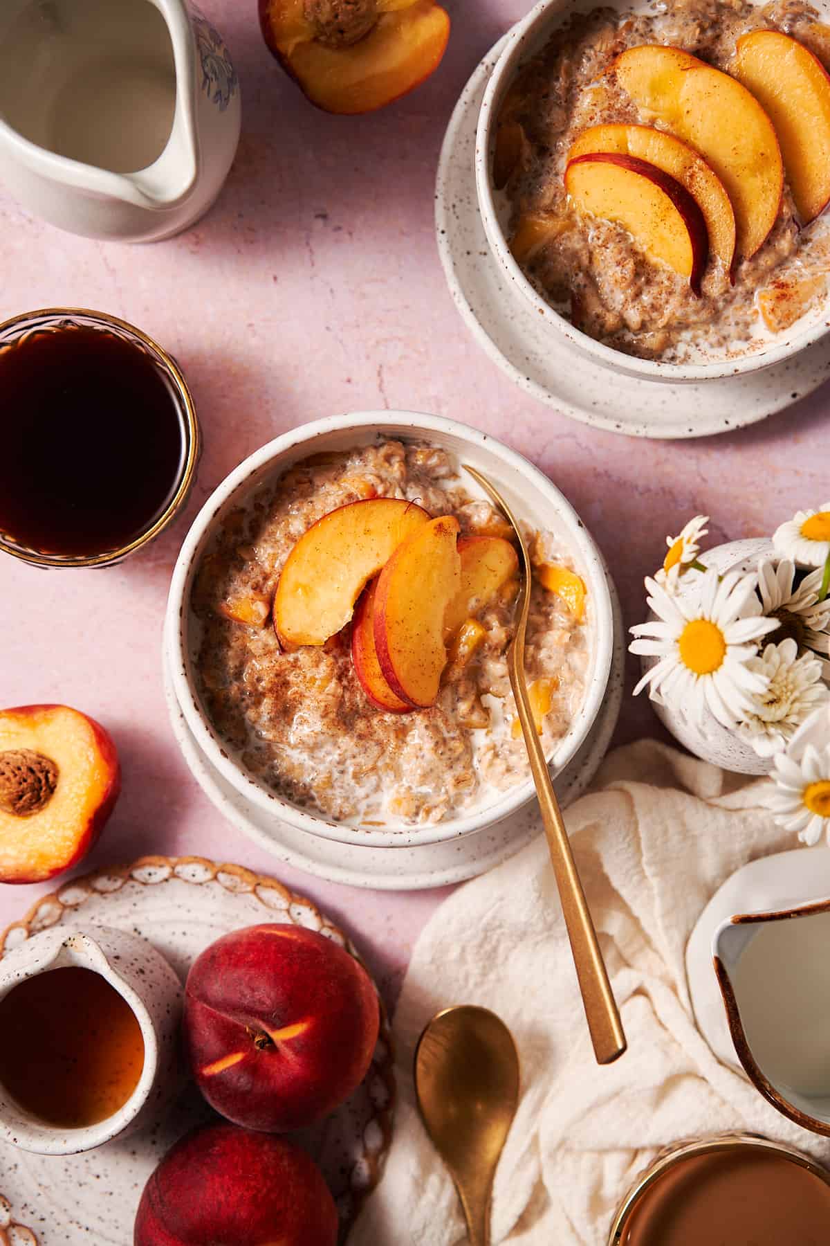 flat lay shot of peaches and cream oatmeal, with daisies in a ceramic vase, fresh coffee, maple syrup, and fresh peaches on the scene.