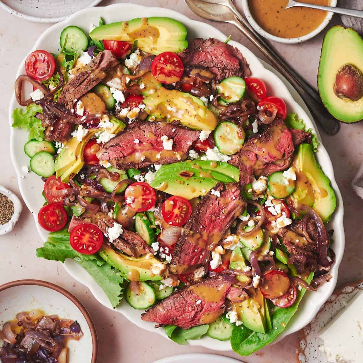 flat lay shot of a vibrant steak salad with tomatoes, avocado, grilled onions, feta, and dressing with grilled bread on the side.