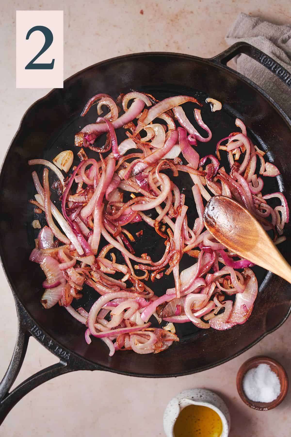 cooked and softened red onion strips in a cast iron skillet with a wooden spoon in the skillet. 