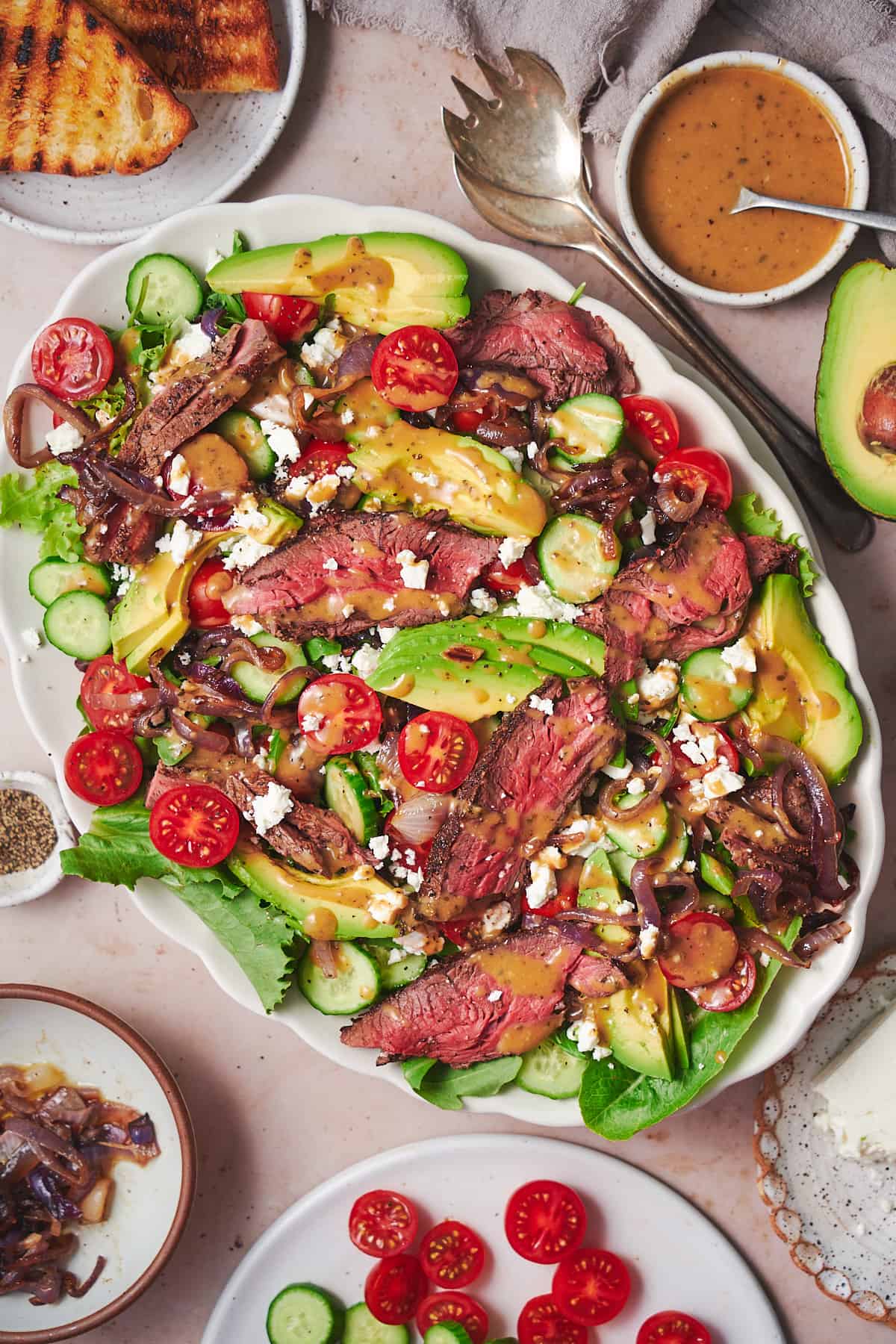flat lay shot of a vibrant steak salad with tomatoes, avocado, grilled onions, feta, and dressing with grilled bread on the side. 