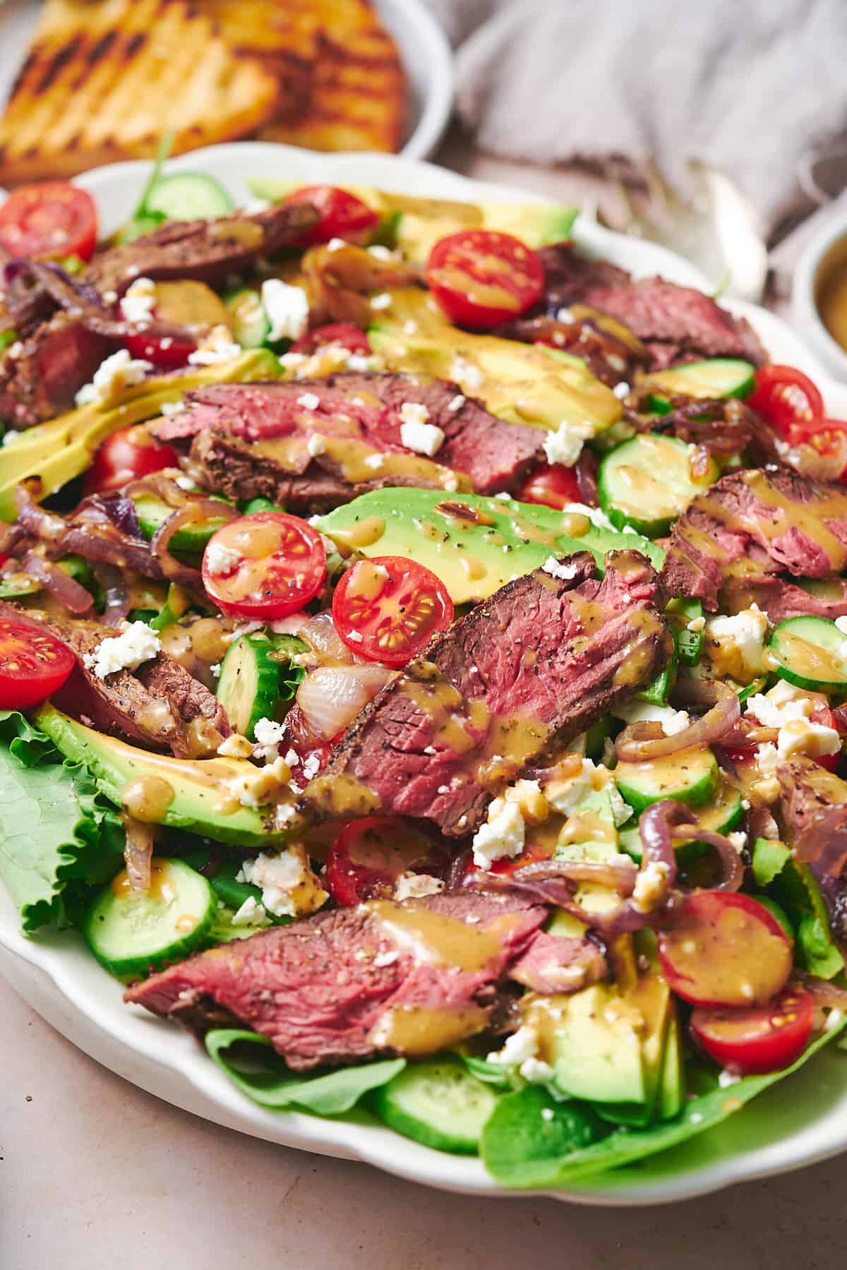 45-degree photo of a steak salad on a ruffled serving plate with avocado, tomato, cucumbers, feta, onions, and dressing. 