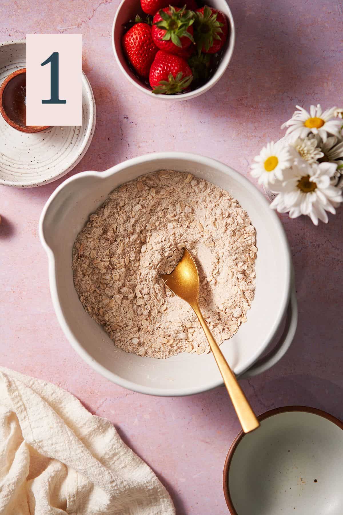 dry ingredients of flour, oats, cinnamon, sugar, and salt in a mixing bowl surrounded by flowers and strawberries. 