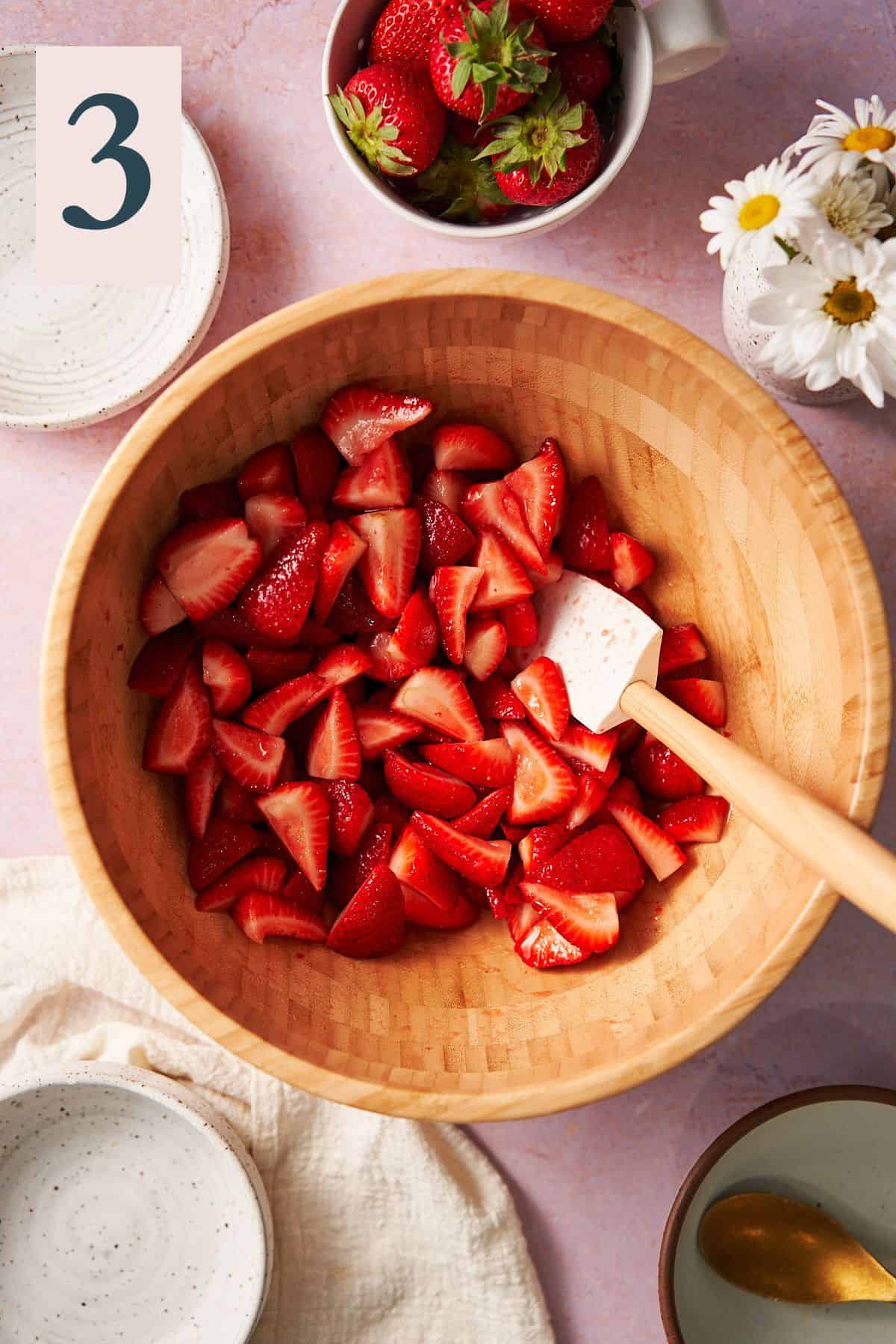 strawberries in a large wooden mixing bowl with a rubber spatula, surrounded by daisies and fresh strawberries. 