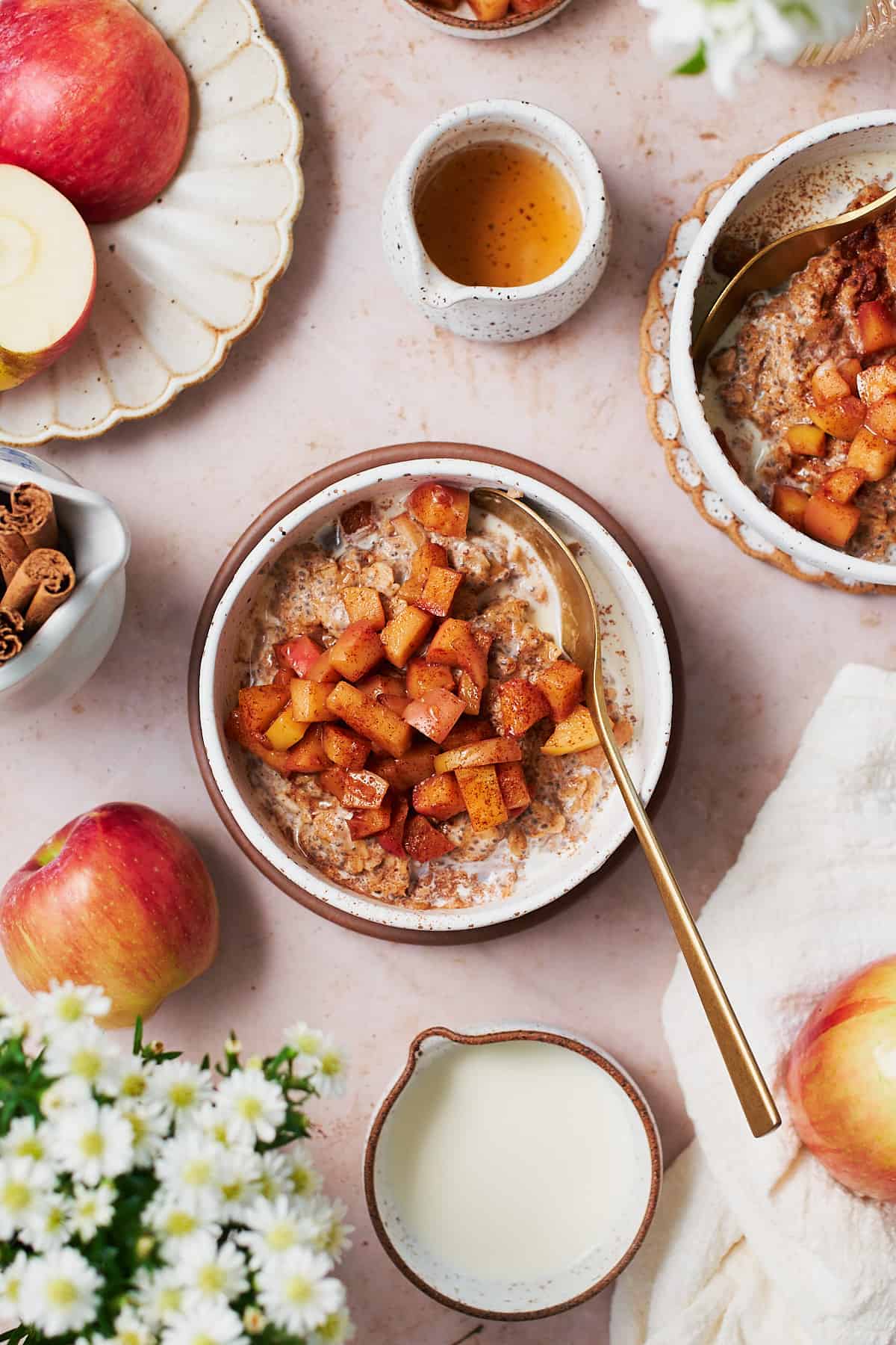 overhead view of apple cinnamon oatmeal in bowls, surrounded by small white flowers, apples, cinnamon sticks, maple syrup a cream.