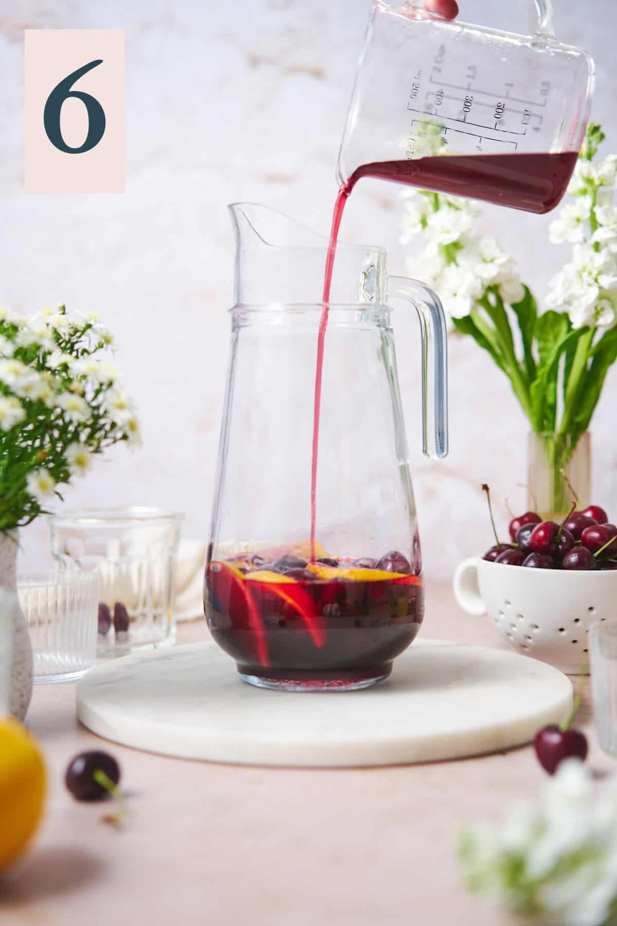 Hand pouring cherry simple syrup into a large pitcher with lemon slices and cherries, with white flowers in the background. 