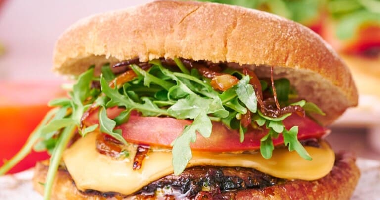 straight on shot of a juicy portobello mushroom burger with arugula, melty cheese, tomatoes, and caramelized onions.
