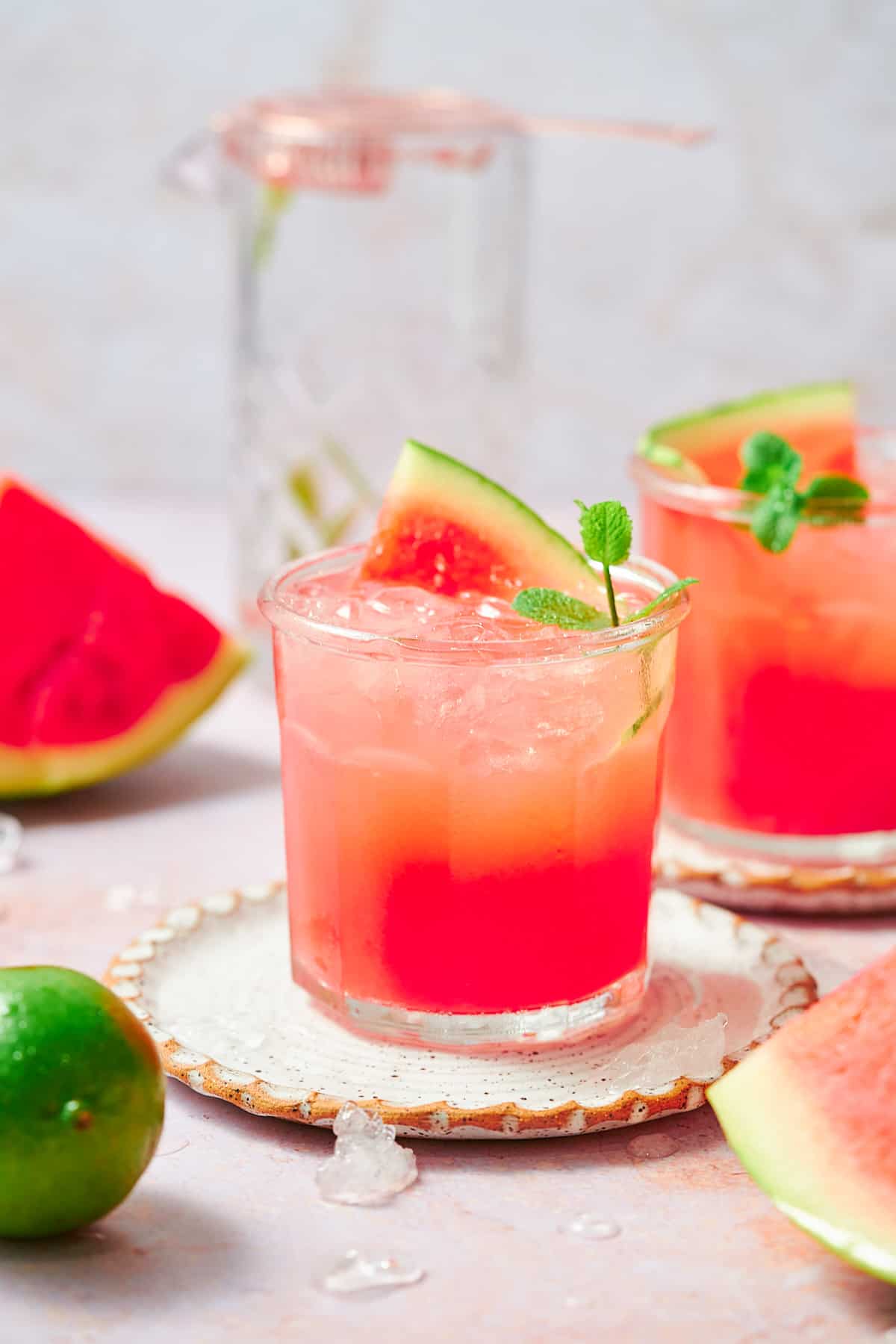 straight on shot of two watermelon vodka cocktails on small ruffled plates, with a mixing glass in the background and watermelon pieces in front. 