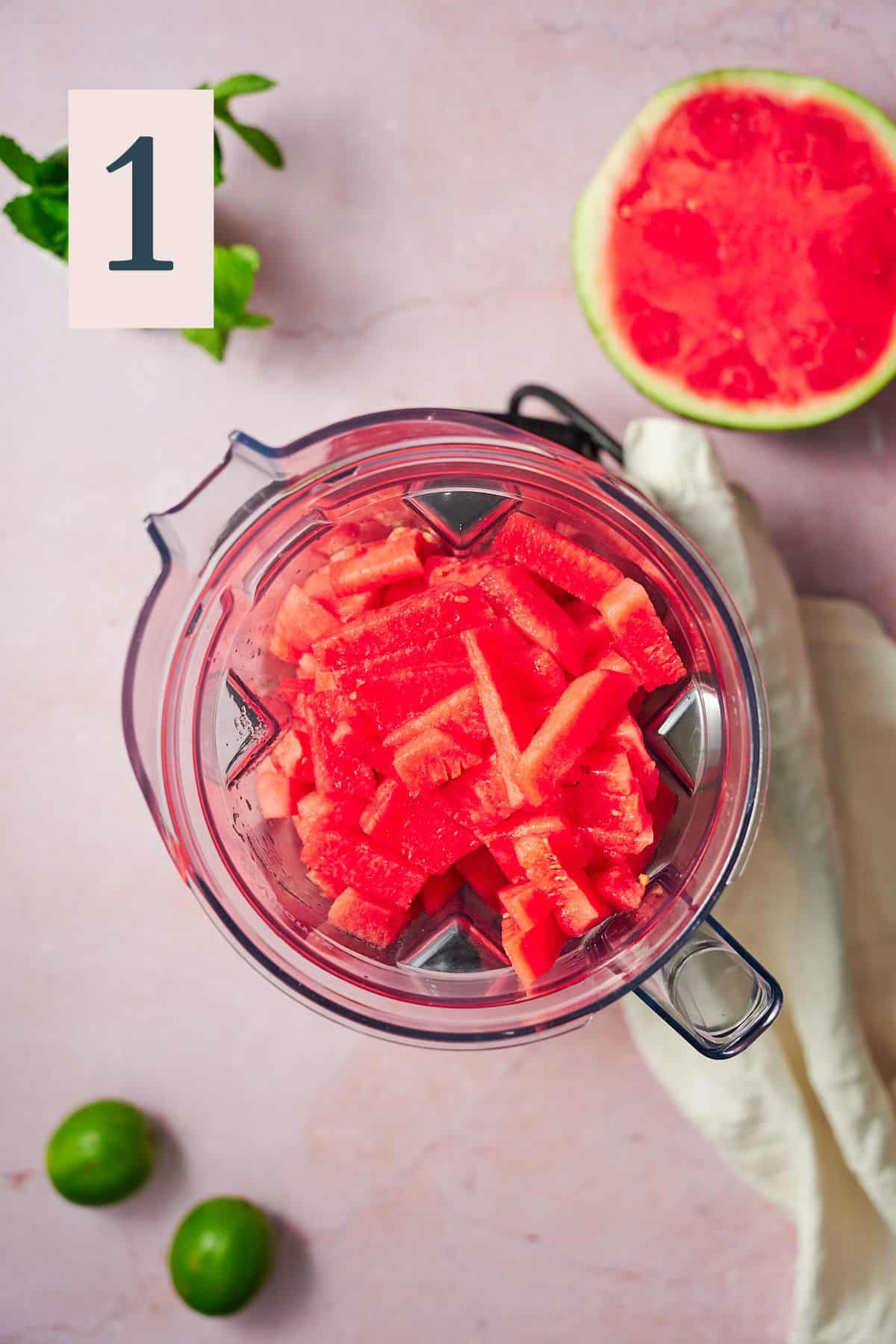 watermelon chunks in a blender, with half a watermelon on the table beneath it. 
