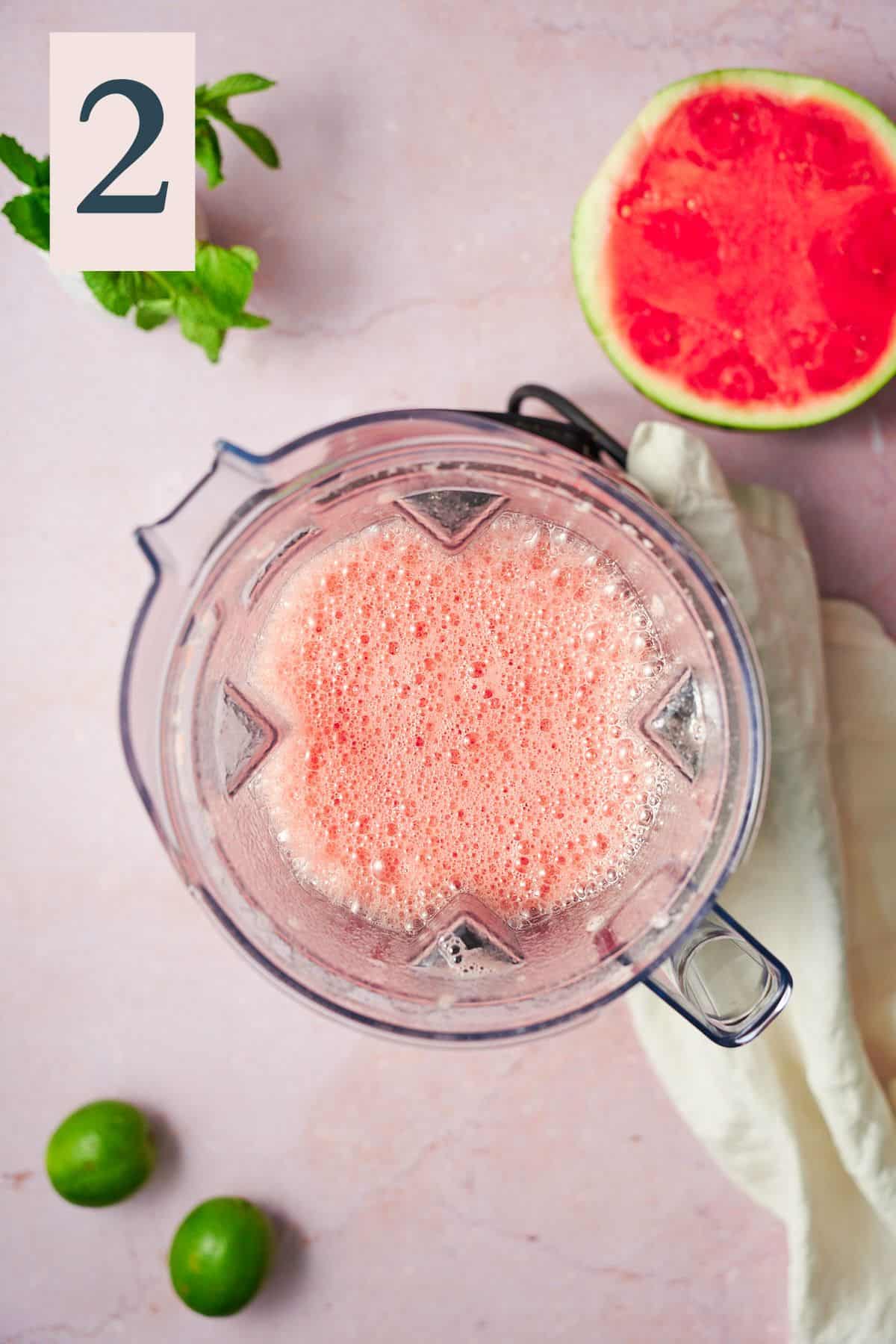 blended watermelon juice with a foamy top in a blender, with a half of a watermelon beneath it. 
