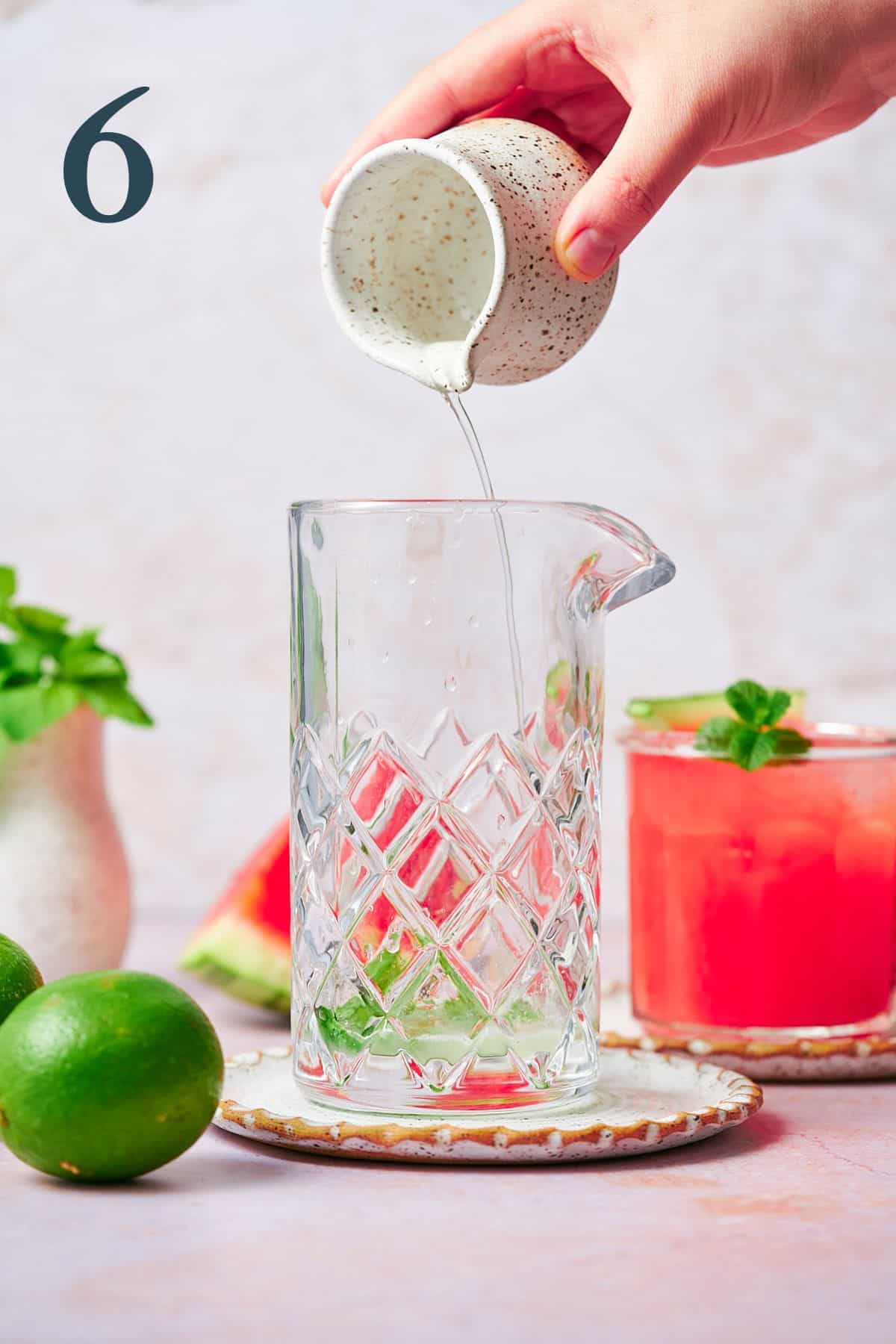 hand pouring simple syrup into the bottom of a mixing glass with a watermelon cocktail in the background. 