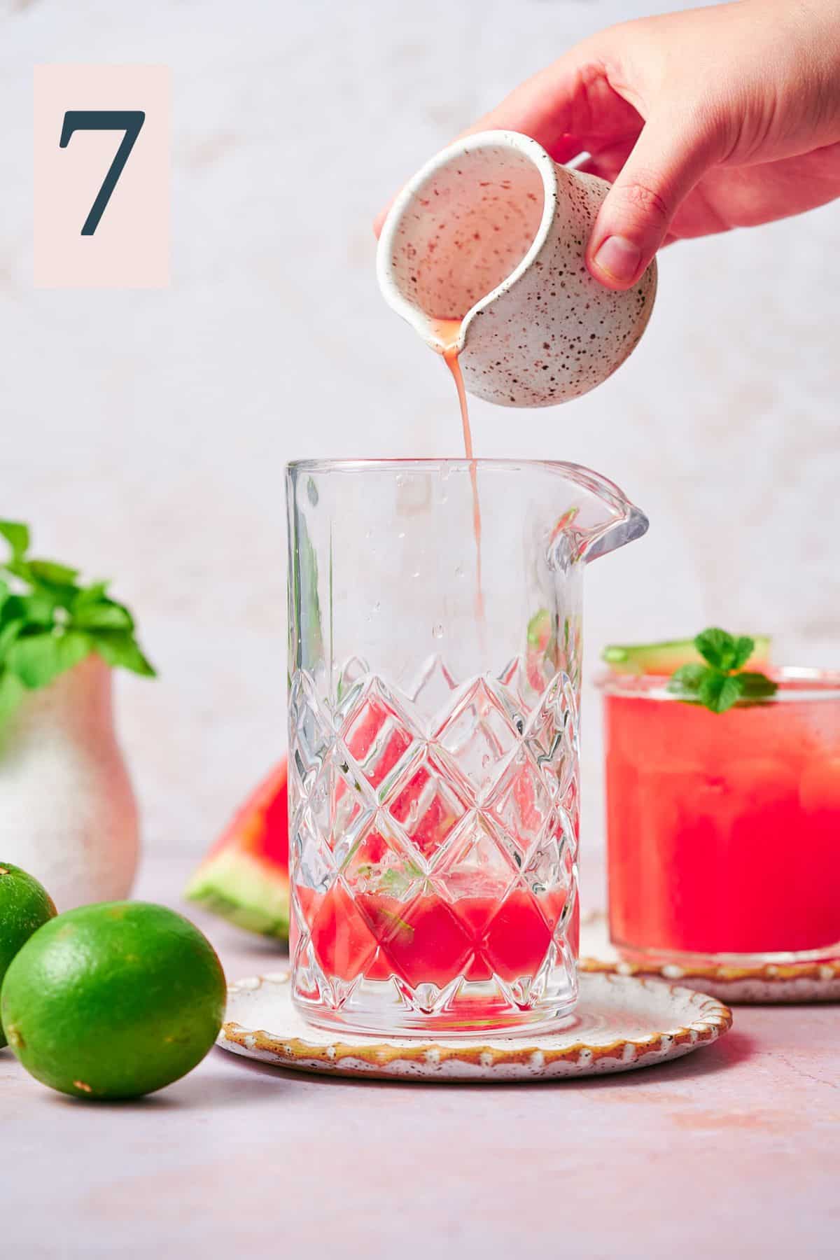 hand pouring watermelon juice into the bottom of a mixing glass with a watermelon cocktail in the background. 