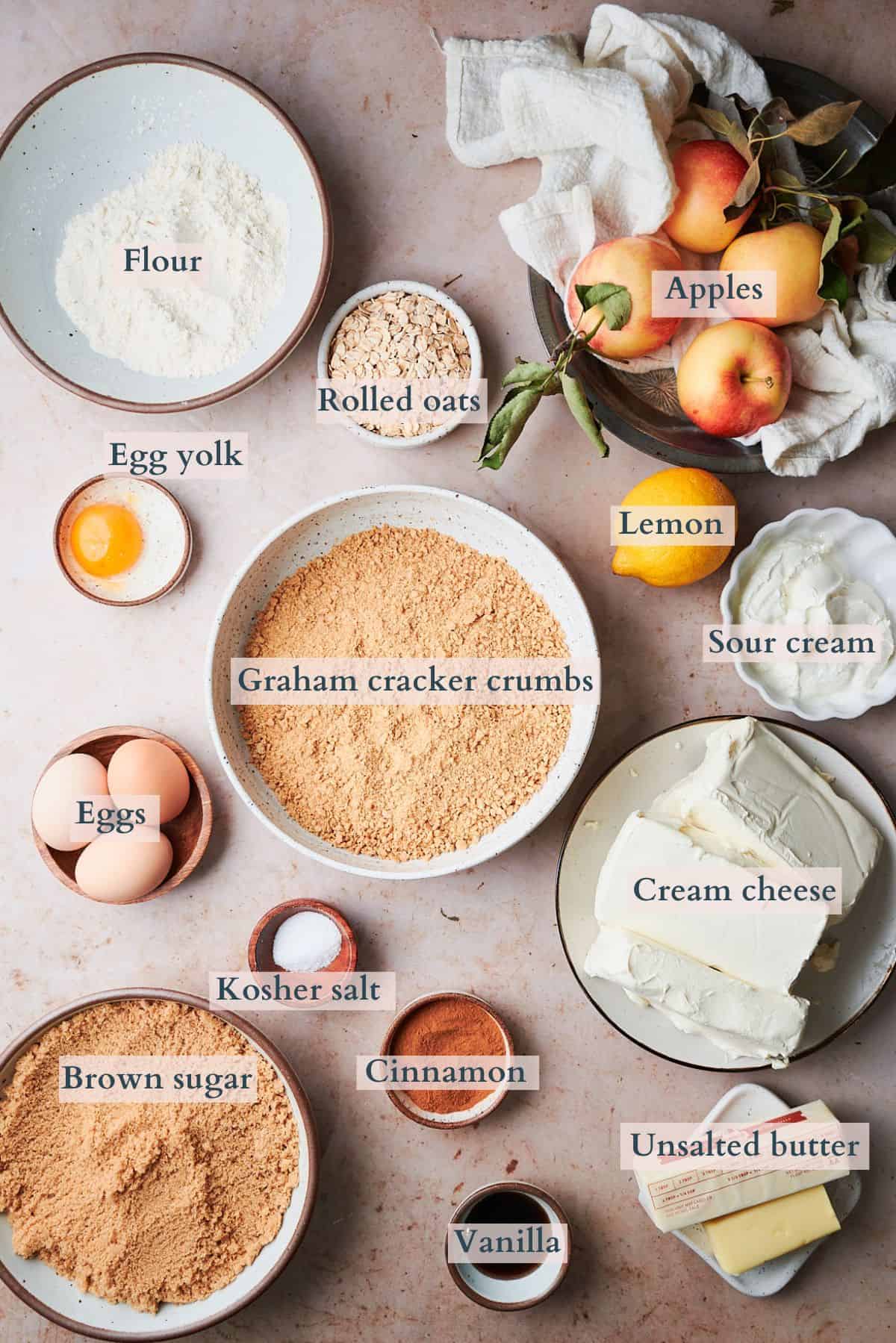 Ingredients to make an apple crisp cheesecake laid out in small bowls and labeled to denote each ingredient. 