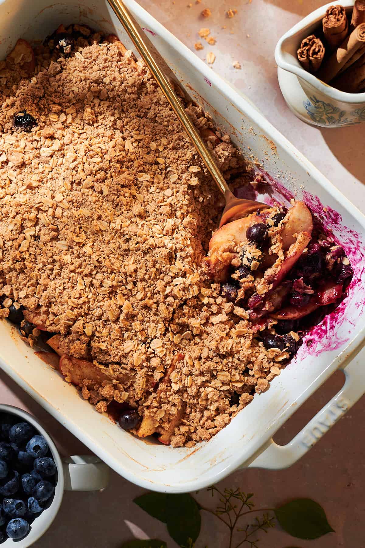Apple and blueberry crumble in a baking dish with a spoon showing off the inside underneath the crumbly oat topping. 