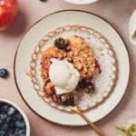 Blueberry and apple crumble on a ruffled plate topped with vanilla ice cream surrounded by ingredients used to make the crumble and a square baking dish with crumble in it above it.
