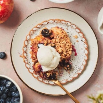 Blueberry and apple crumble on a ruffled plate topped with vanilla ice cream surrounded by ingredients used to make the crumble and a square baking dish with crumble in it above it.