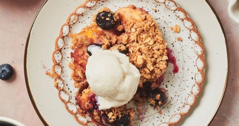 Blueberry and apple crumble on a ruffled plate topped with vanilla ice cream surrounded by ingredients used to make the crumble and a square baking dish with crumble in it above it.