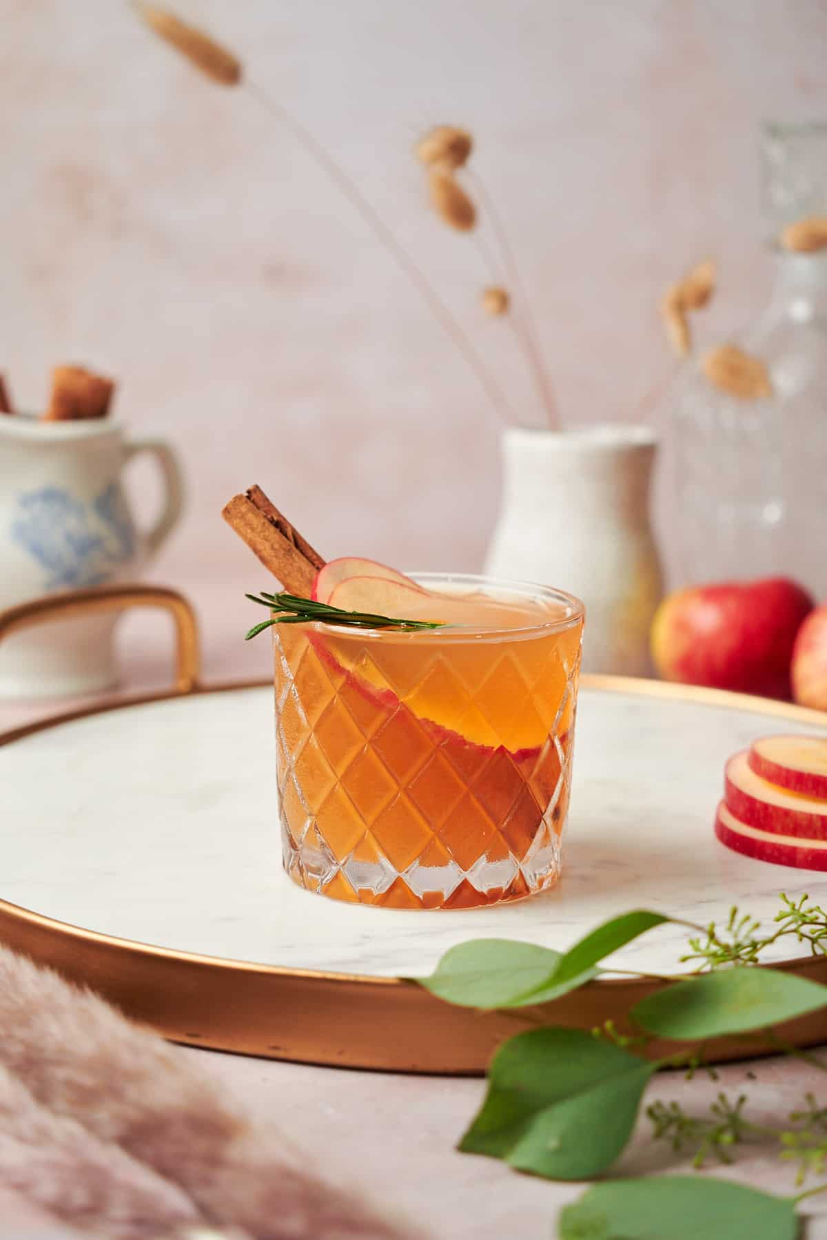 photo of an apple cider bourbon cocktail with a cinnamon stick and rosemary sprig, with apples and pampas grass in the background.