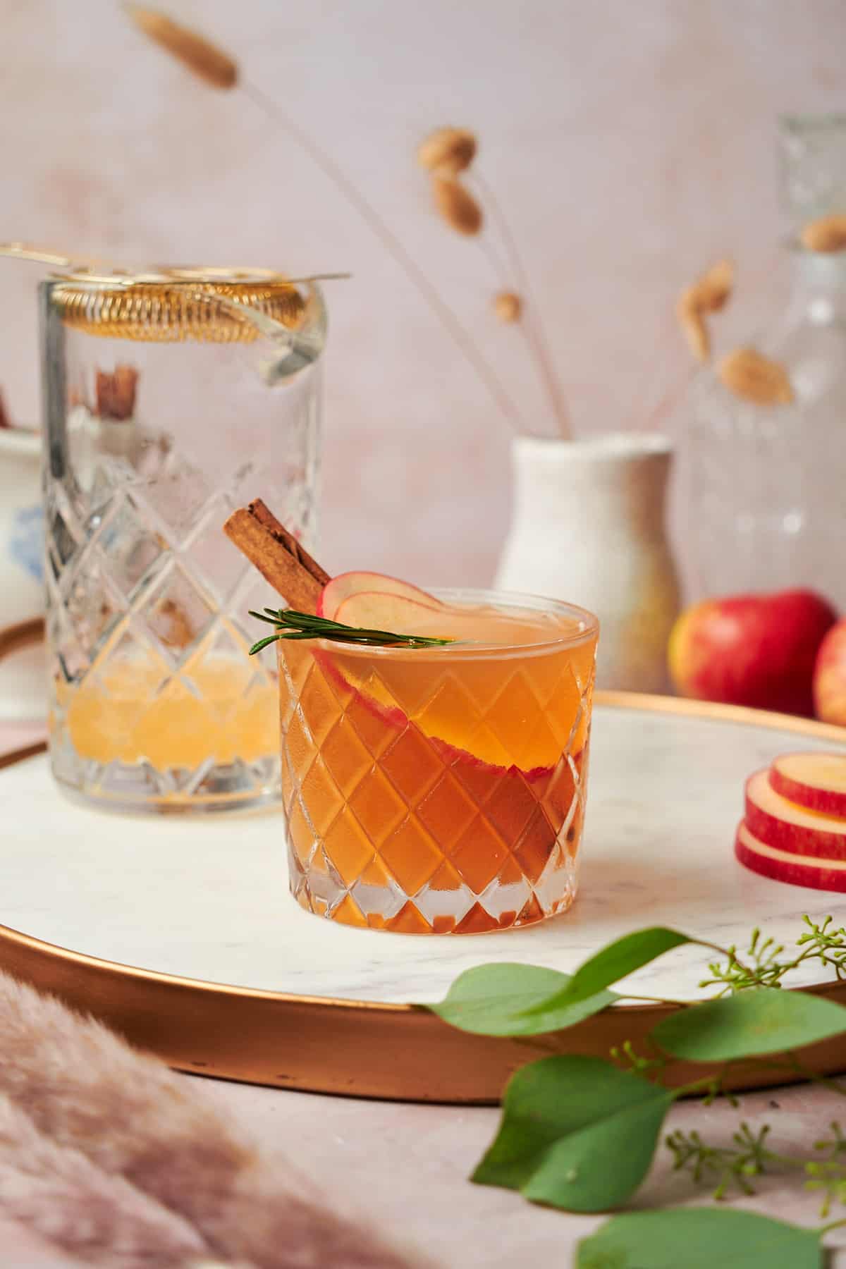 photo of an apple cider bourbon cocktail with a cinnamon stick and rosemary sprig, with apples, pampas grass, and a mixing glass in the background. 