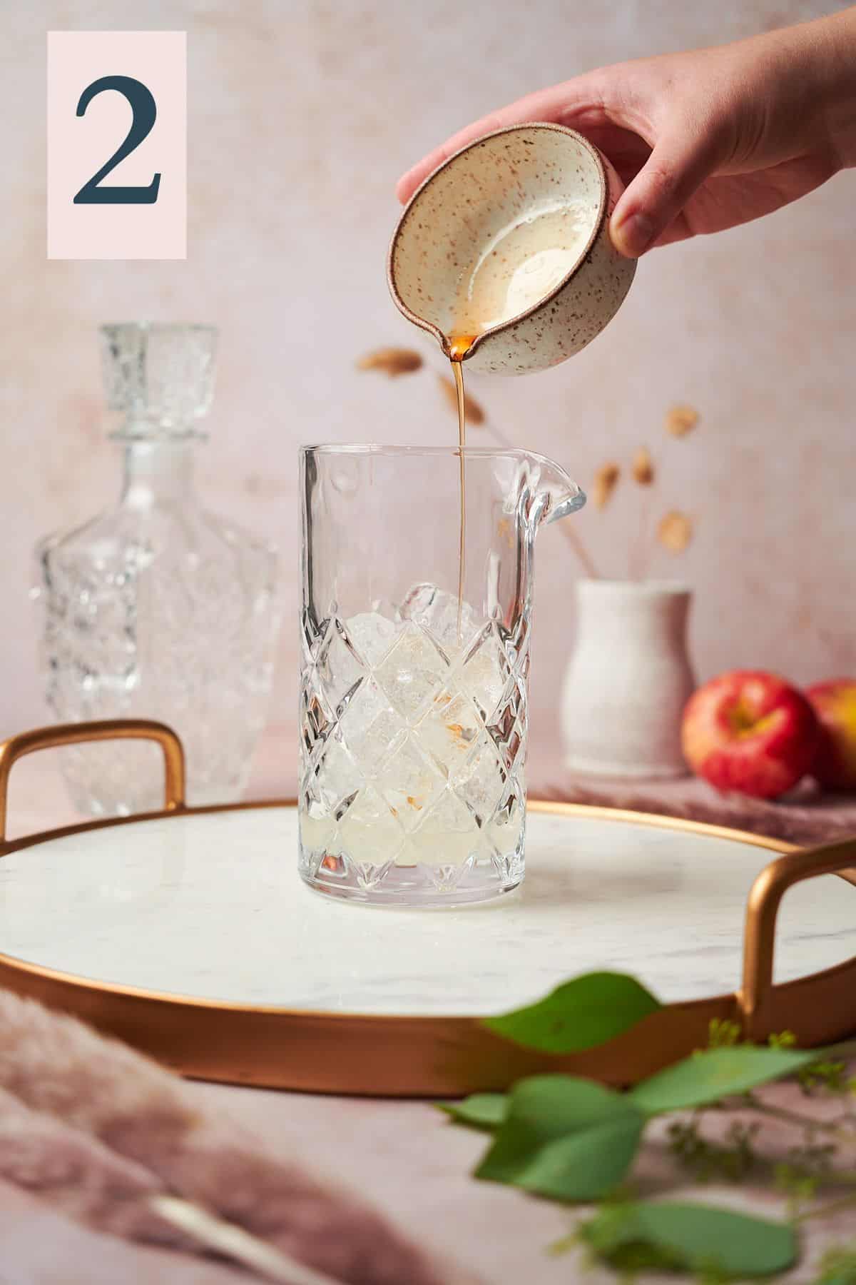 Hand pouring maple syrup into a mixing glass with pampas grass and apples in the background. 
