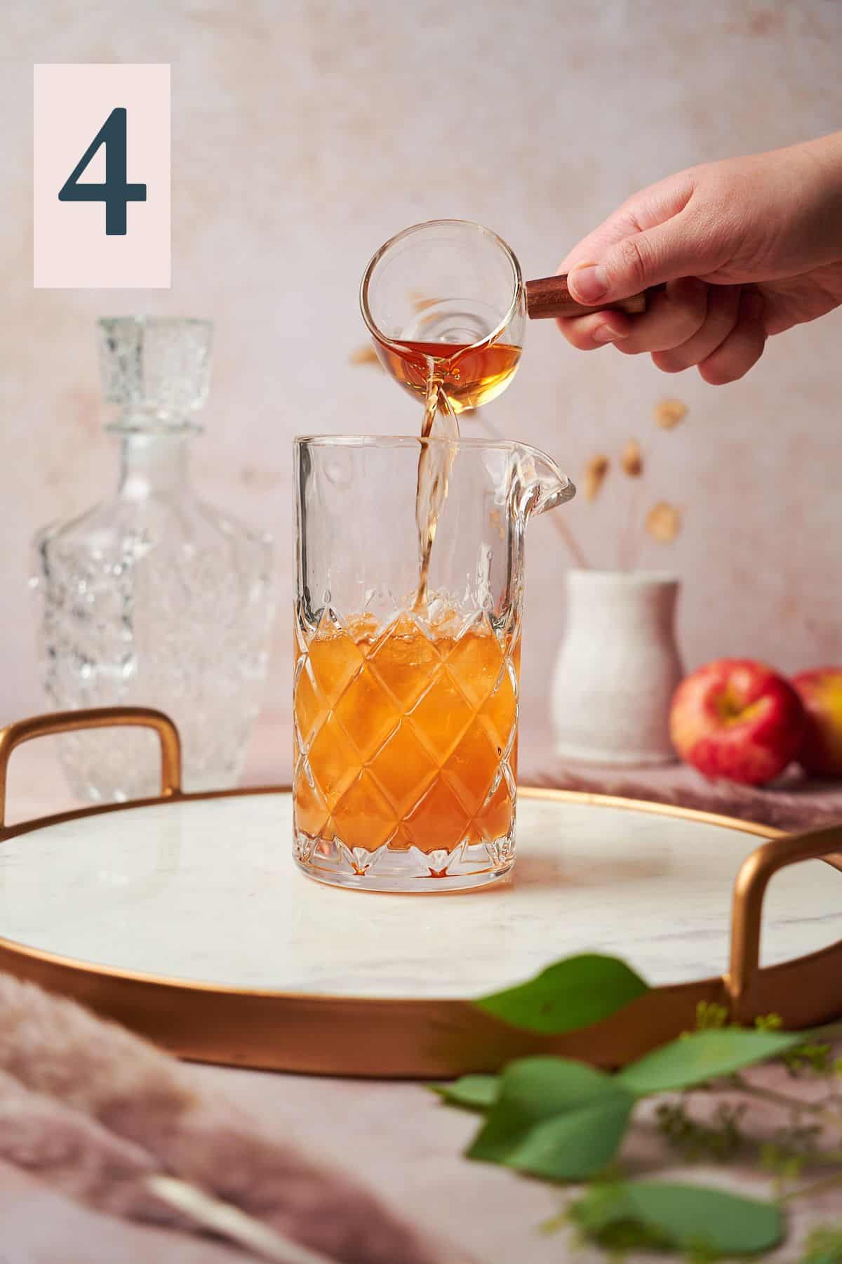 Hand pouring bourbon into a mixing glass with pampas grass and apples in the background.