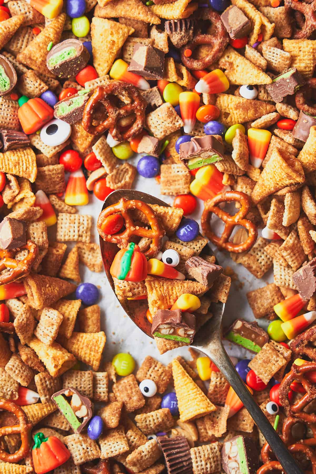 Snack mix with Halloween candy, candy eyes, pretzels, bugles, and candy corn o na baking sheet in a large scooping spoon. 