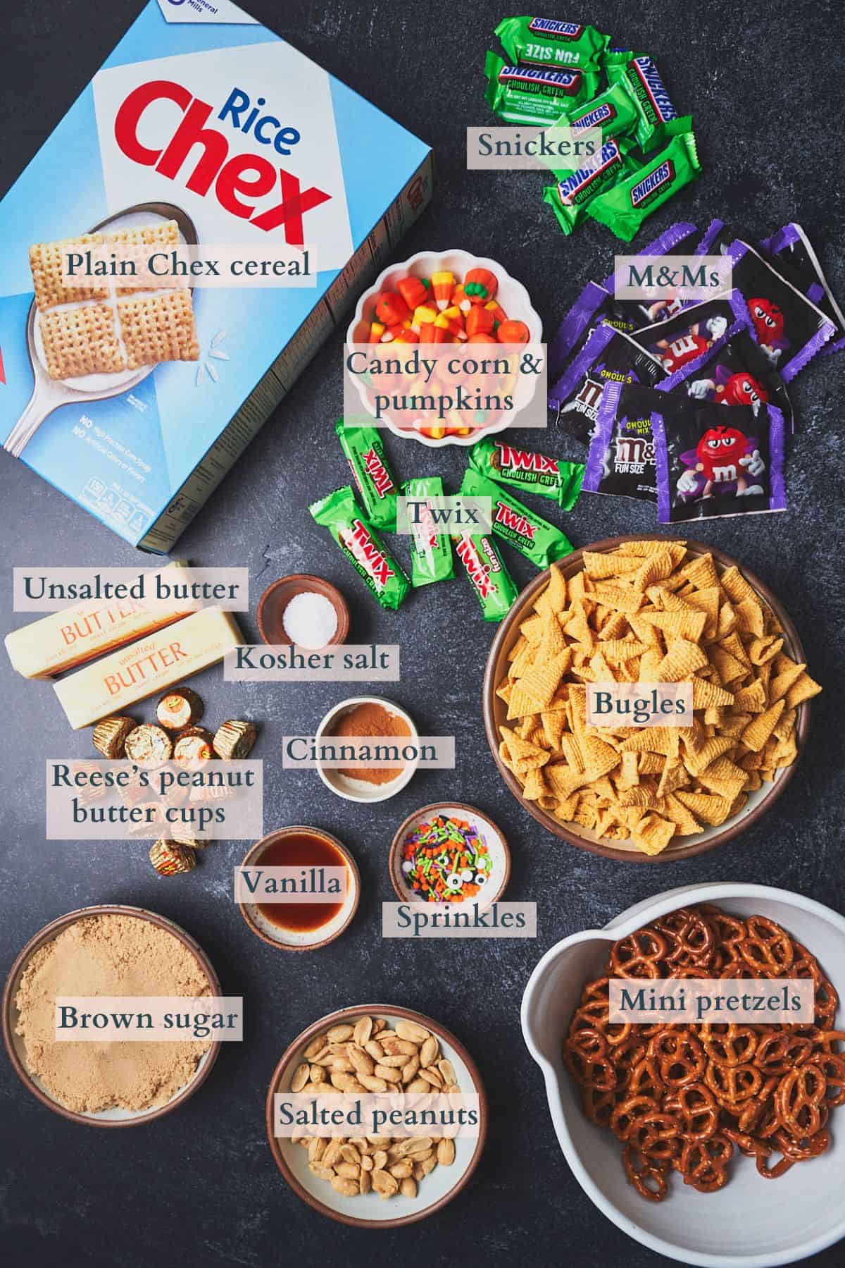 Ingredients to make a sweet and salty Halloween snack mix laid out on a dark backdrop in small bowls and labeled to denote each ingredient. 