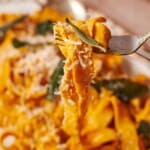 Fork full of pumpkin pasta with pappardelle pasta and topped with grated parmesan cheese and fried sage.