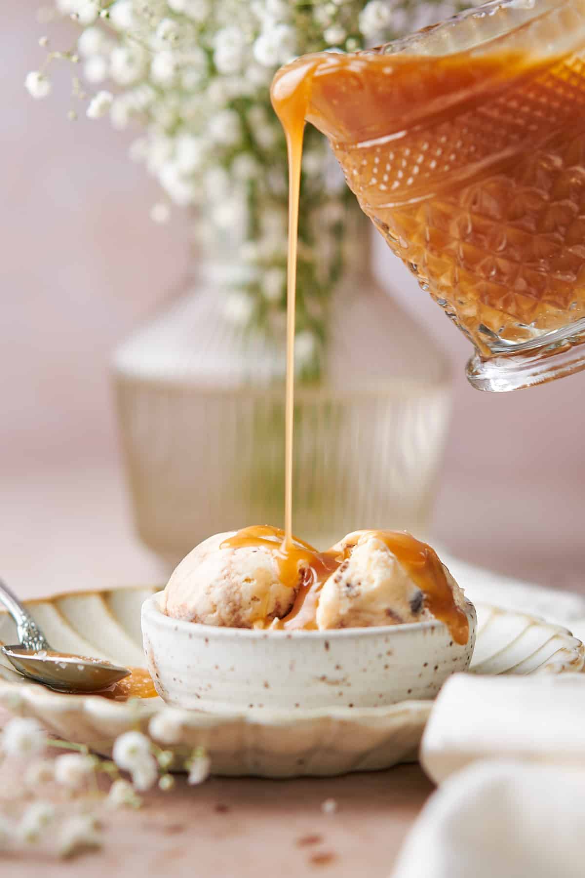 hand pouring salted caramel from a glass container onto ice cream in a small bowl on a ruffled plate with baby's breath flowers in the background. 