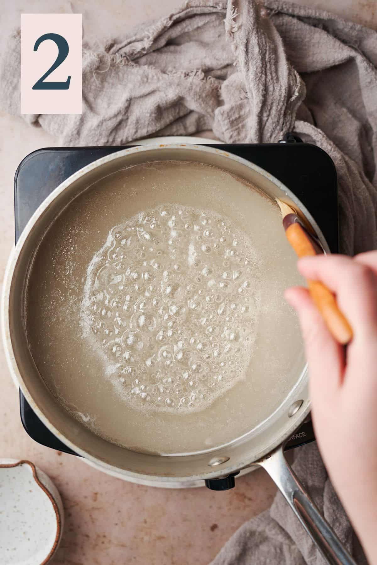 hand using a pastry brush to brush around the sides of a bubbling sugar water mixture in a saucepan. 