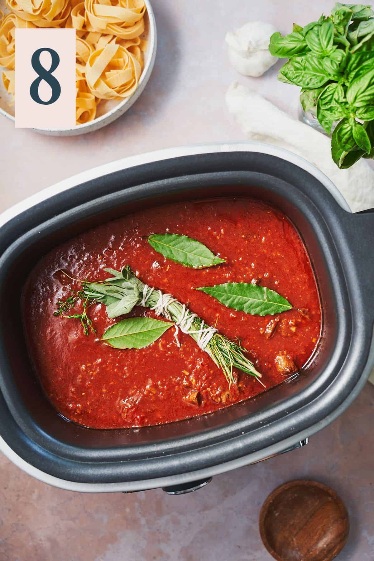 Tomato sauce topped with bay leaves and an herb bundle in a slow cooker. 