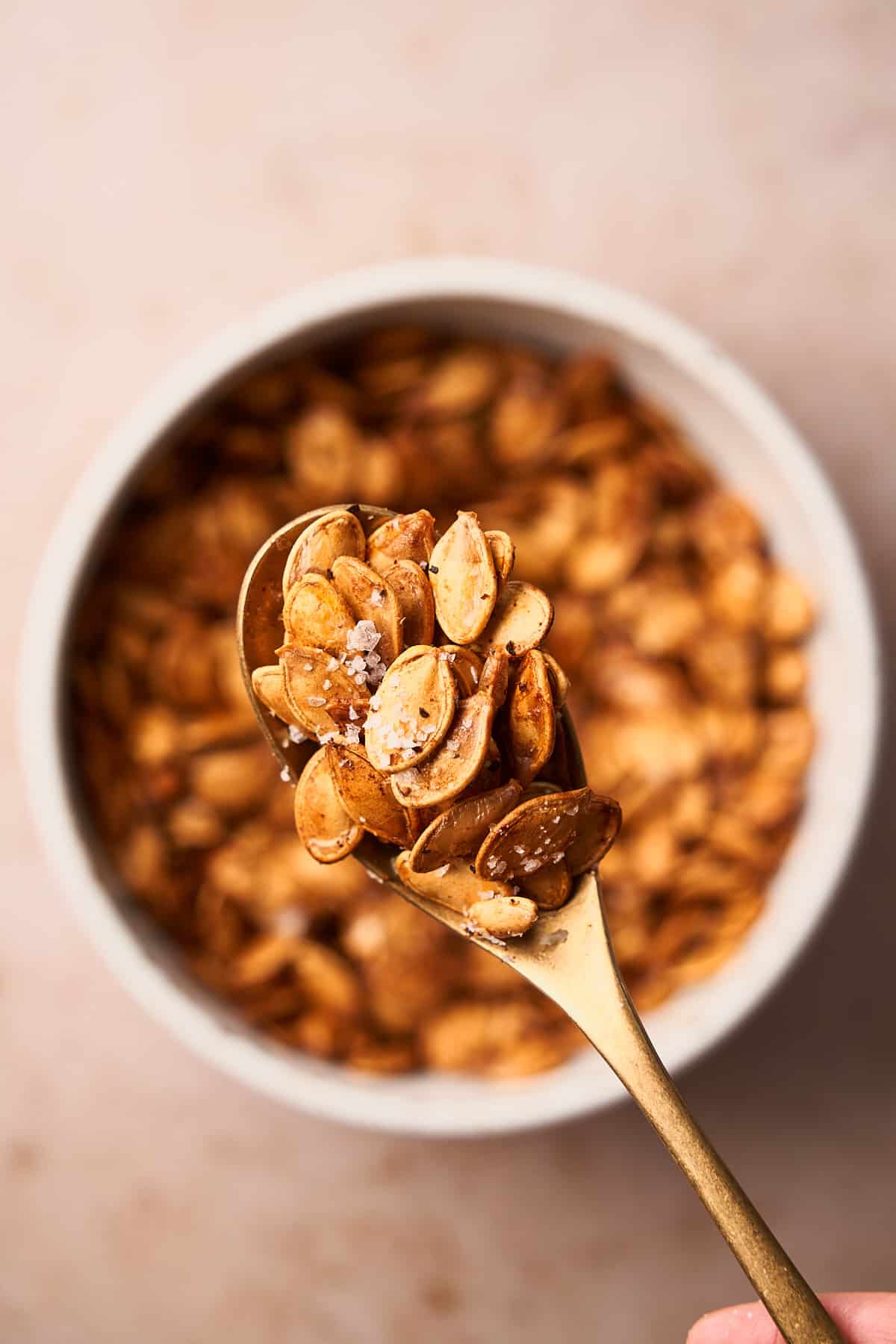 Salted air fryer pumpkin seeds on a golden spoon, held over a bowl of seeds.