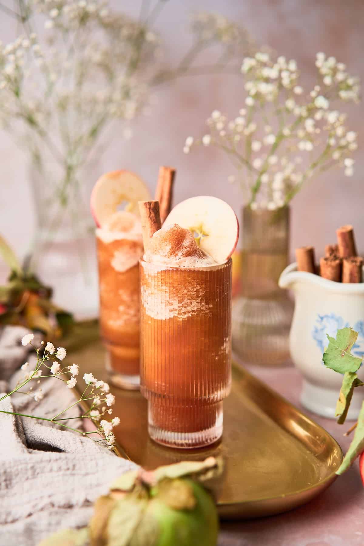 Straight on shot of an apple cider slushie in a ribbed glass, served with a cinnamon stick and an apple round in the glass, with baby's breath flowers surrounding it. 
