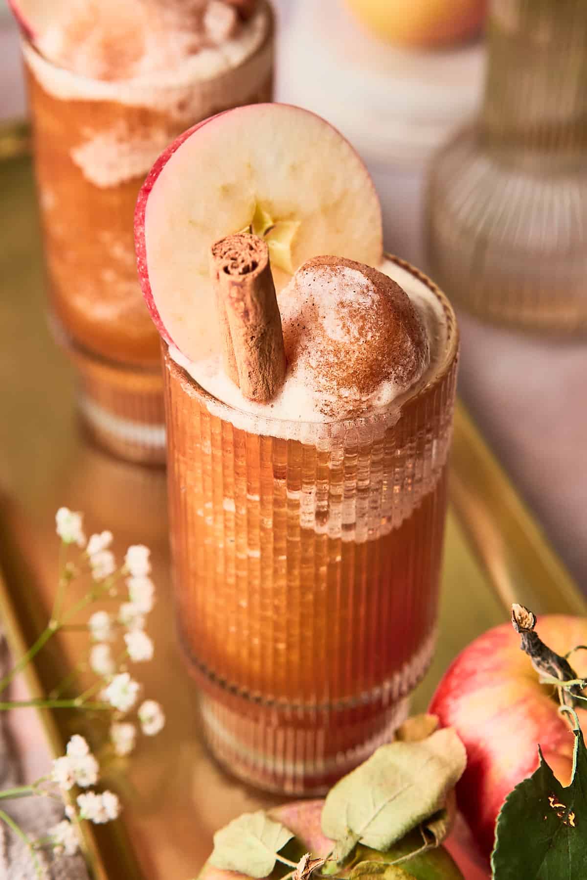 45-degree shot of ab apple cider slushie with a cinnamon stick, and an apple slice on a golden tray with baby's breath flowers and an apple with leaves still attached. 