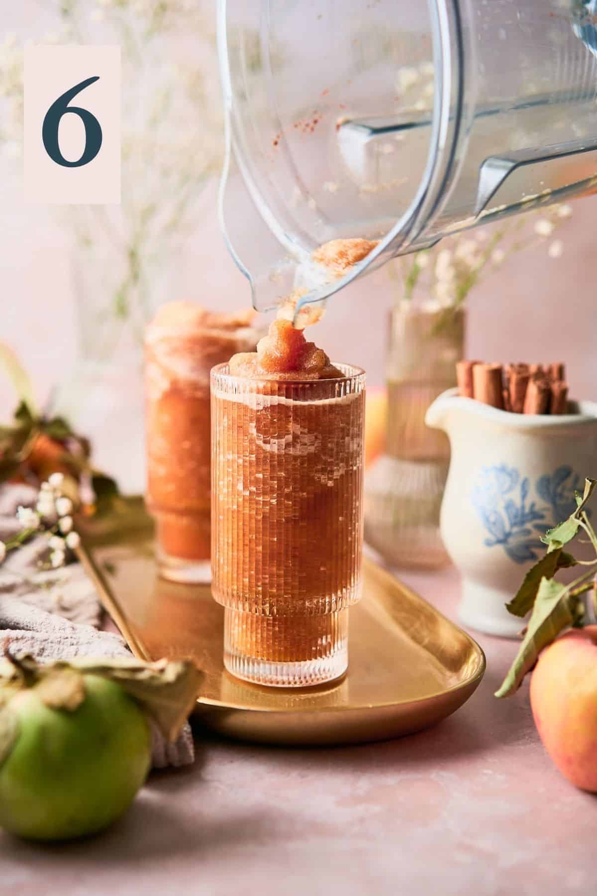 pouring blended apple cider into serving cups,, surrounded by cinnamon ticks, flowers, and fresh apples. 