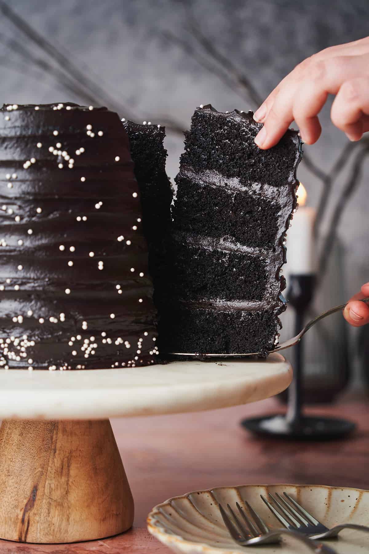 Hand pulling a 4-layer slice of black velvet cake from the cake with a candlestick burning in the background. 
