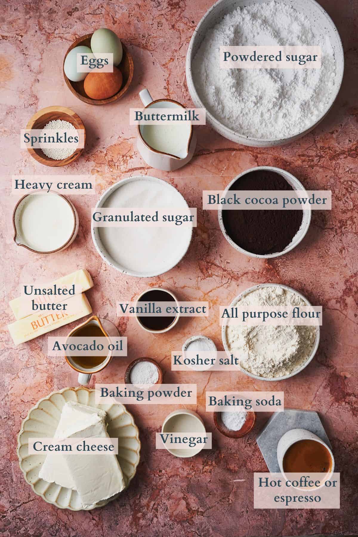 Ingredients to make a black velvet cake laid out on a table in small bowls and on plates with text overlaying to denote each ingredient. 