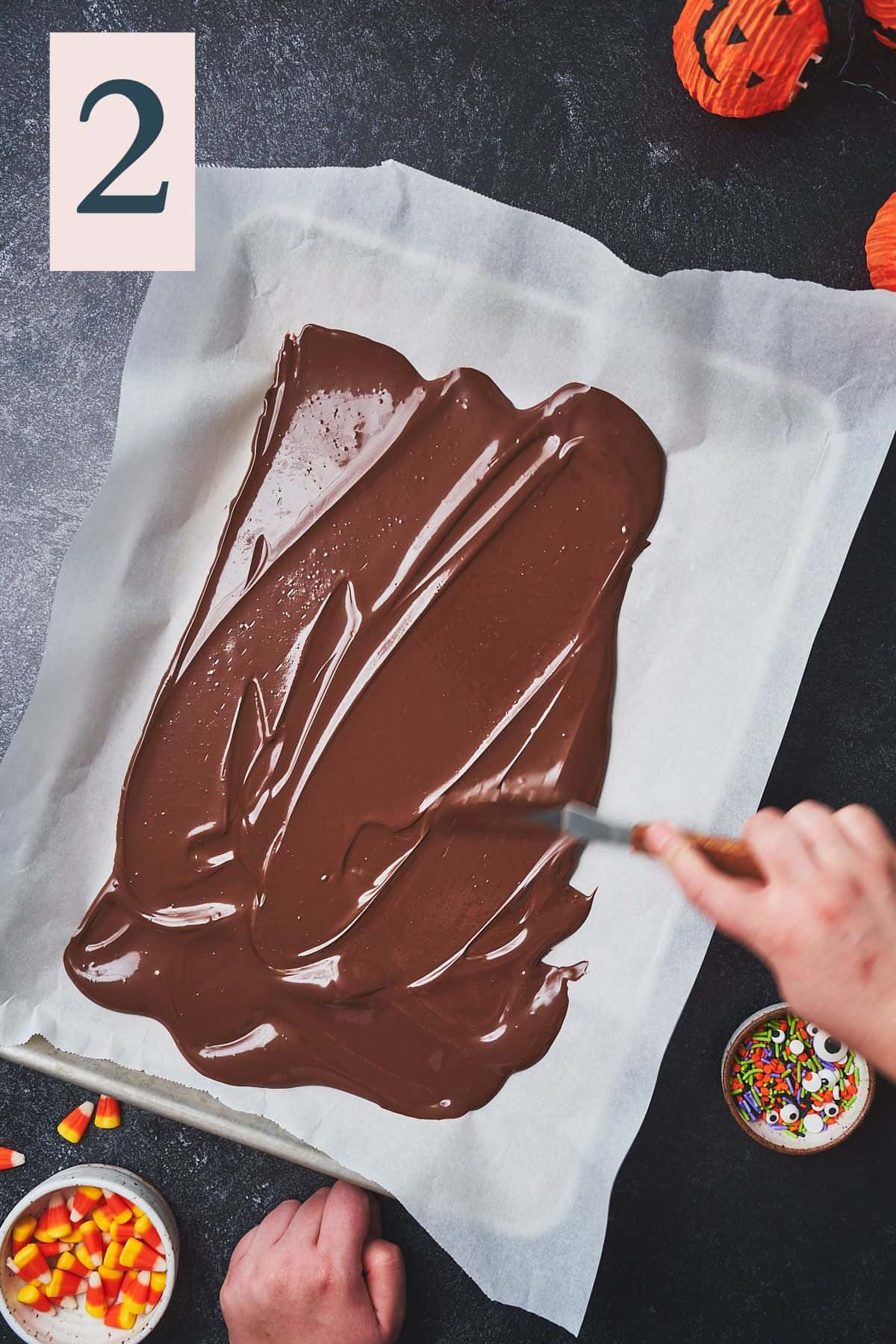 hand spreading melted dark chocolate with an offset spatula on a parchment lined baking sheet.